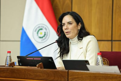 Claudia Centurión, con blusa clara y abrigo neutro, habla en mesa con micrófono, una bandera de Paraguay al fondo.