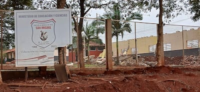 Cartel de la Escuela Básica N° 975 San Miguel, con lema 'Educación es compromiso' y ruinas visibles de un edificio escolar.