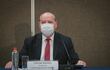 César Diesel sentado en un escenario con mascarilla, traje oscuro y corbata a rayas, rodeado de un ambiente sobrio y profesional.