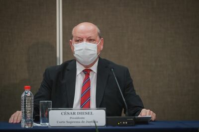 César Diesel sentado en un escenario con mascarilla, traje oscuro y corbata a rayas, rodeado de un ambiente sobrio y profesional.