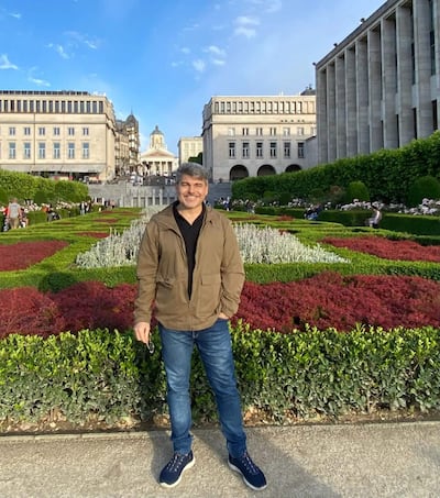 Hombre de pie en un jardín, sonriendo, viste abrigo beige y jeans azules, con plantas verdes y flores alrededor.