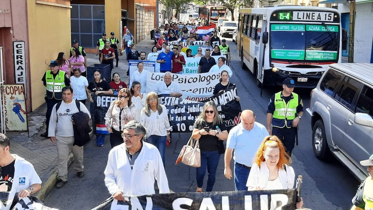 Grupo de médicos en batas blancas marchan con carteles en una calle urbana, rodeados de policías y vehículos.