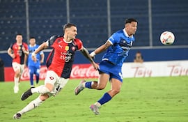 Dos hombres de pie, uno con mochila, conversan en un acceso del estadio, con asientos en rojo, blanco y azul al fondo.