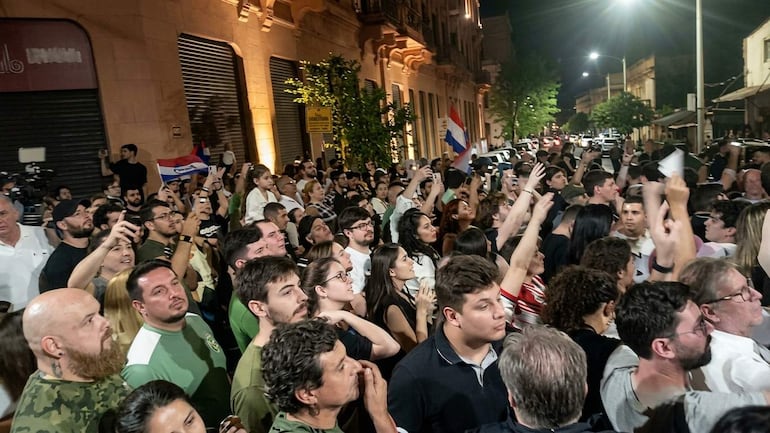 Multitud festiva en la calle, ondeando banderas paraguayas y con expresiones de alegría y tranquilidad.