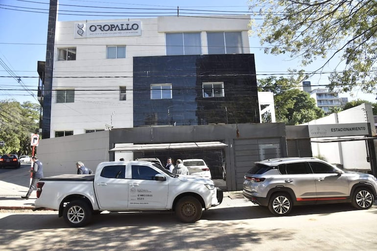 Personas en chaquetas oscuras conversan cerca de un vehículo oficial junto al edificio Oropallo Institute of Genetics.