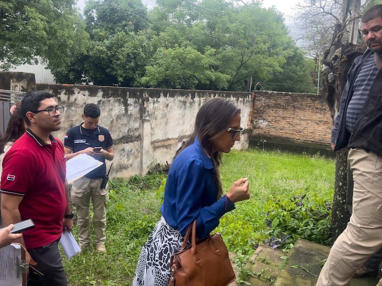 Hombre con gafas y camiseta roja observa mientras una mujer en blusa azul habla con un hombre de chaqueta oscura en un patio.