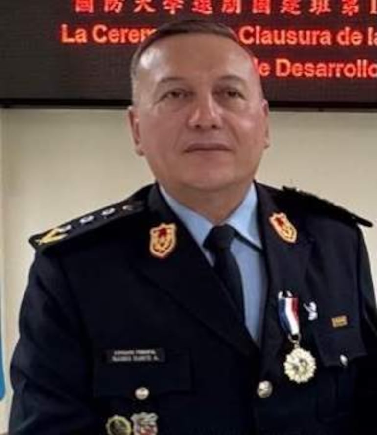 Oficial en uniforme militar con medallas, posando de frente en un ambiente interior iluminado con cartel de ceremonia.