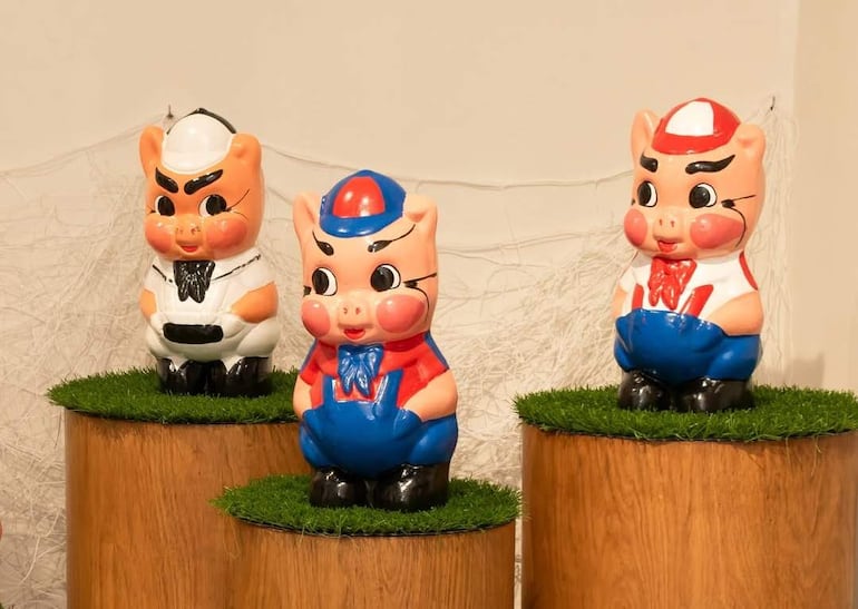 Tres cerditos de cerámica en colores variados, expuestos en pedestales de madera en un fondo claro simulado césped.