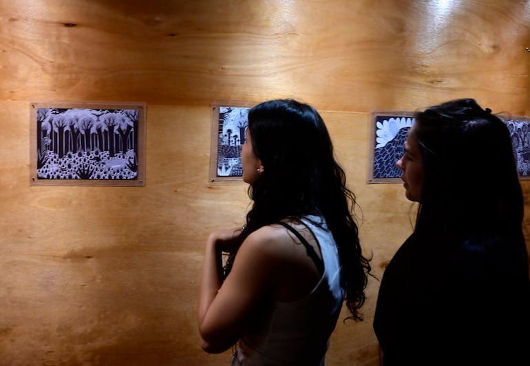 Dos mujeres de pie en galería de arte, una con camiseta blanca y otra con chaqueta negra, observan cuadros en panel de madera.