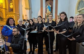 Directora en bata azul dirige coro de mujeres con partituras en iglesia decorada con estatuas religiosas.