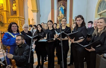 Directora en bata azul dirige coro de mujeres con partituras en iglesia decorada con estatuas religiosas.