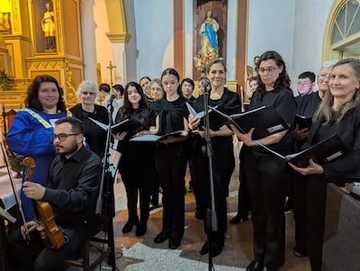 Directora en bata azul dirige coro de mujeres con partituras en iglesia decorada con estatuas religiosas.
