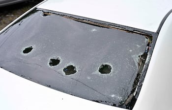 Automóvil blanco dañado con vidrio trasero destrozado, rodeado de fragmentos; entorno urbano de desolación.