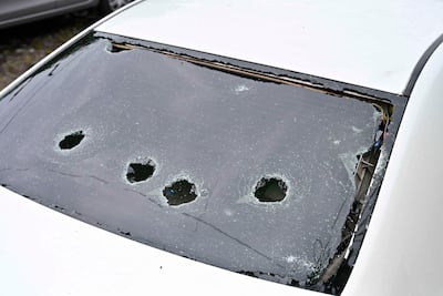 Automóvil blanco dañado con vidrio trasero destrozado, rodeado de fragmentos; entorno urbano de desolación.