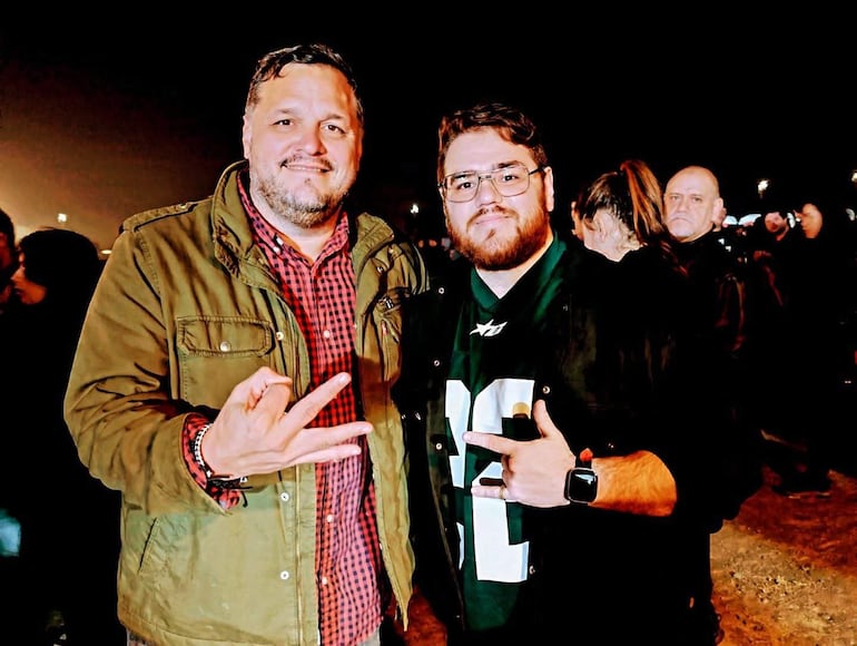 Hombres sonriendo en un evento social, uno en chaqueta verde y el otro en camiseta verde con el número 22.