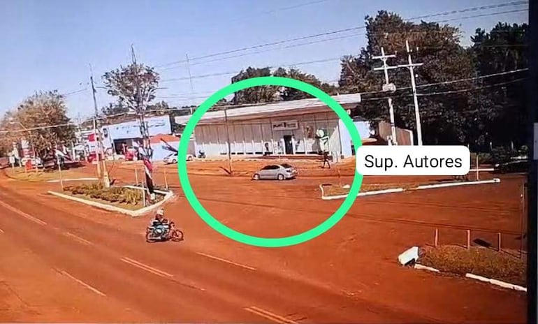 Motociclista circulando por calle de tierra roja, con vehículo gris en la misma vía. Día soleado en escena urbana.
