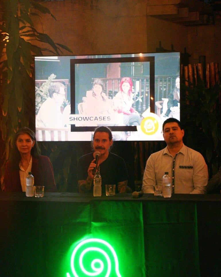 Orador central con micrófono y tatuajes, flanqueado por tres panelistas en un ambiente informal, con decoración colorida y una pantalla detrás.