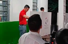 Hombre de camiseta roja votando en un cubículo electoral, rodeado de personas en un ambiente interior de colores claros.