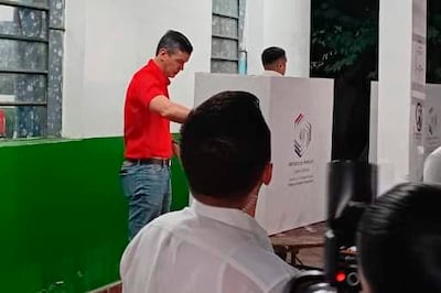 Hombre de camiseta roja votando en un cubículo electoral, rodeado de personas en un ambiente interior de colores claros.