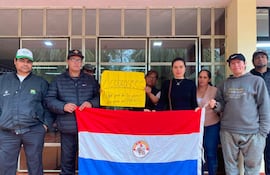 Mujer con expresión seria sostiene una bandera de Paraguay, rodeada de hombres con chaquetas. Cartel amarillo pregunta sobre los salarios.