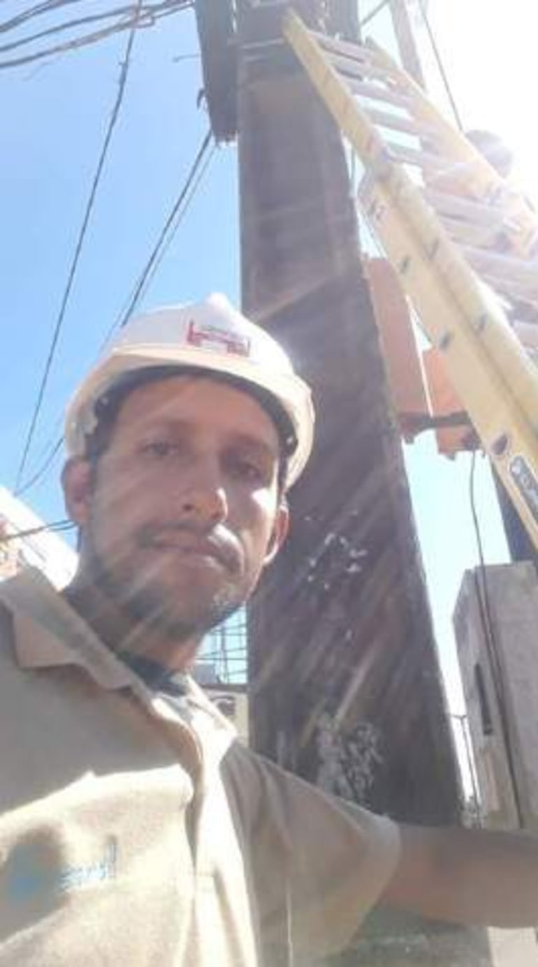 Hombre con casco blanco y camiseta clara sostiene un poste, rodeado de cables eléctricos en un entorno urbano.