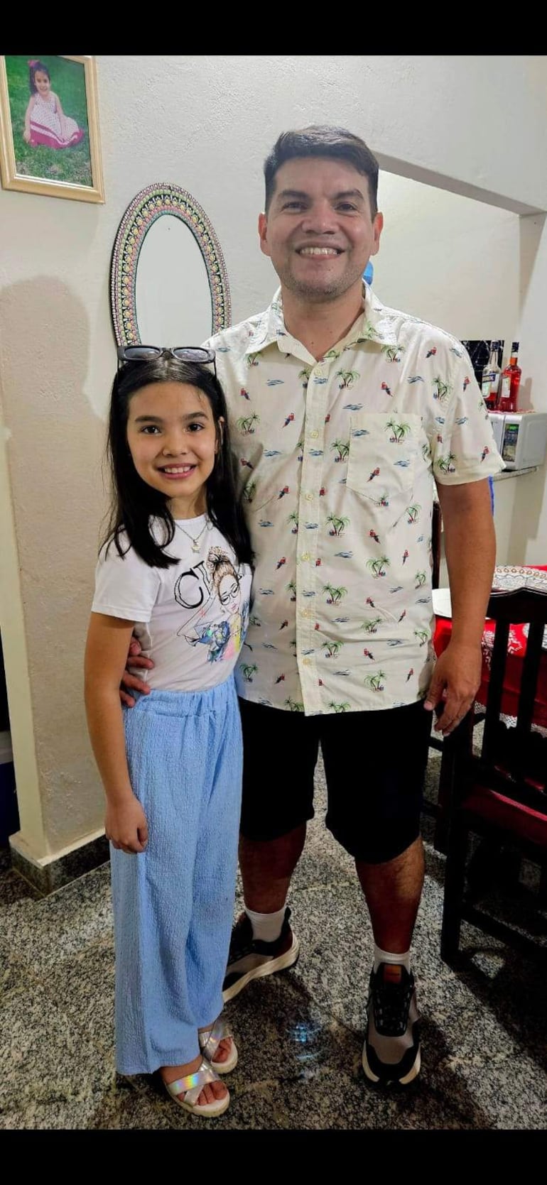 Niña con camiseta blanca y pantalones azules sonríe junto a un adulto en camisa colorida y pantalones oscuros en hogar acogedor.