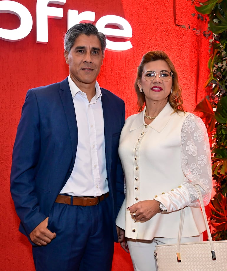 Hombre en traje azul y mujer en blusa blanca sonríen, ambos llevan accesorios formales, fondo rojo y texto decorativo.