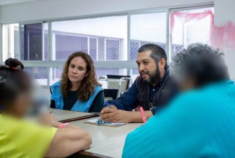 Mujer con cabello rizado y chaleco azul escucha a un hombre con barba y camisa oscura en reunión tranquila.