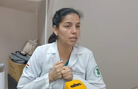 Mujer en uniforme blanco con manos juntas, mostrando interés en conversación, en oficina con objetos cotidianos en la mesa.