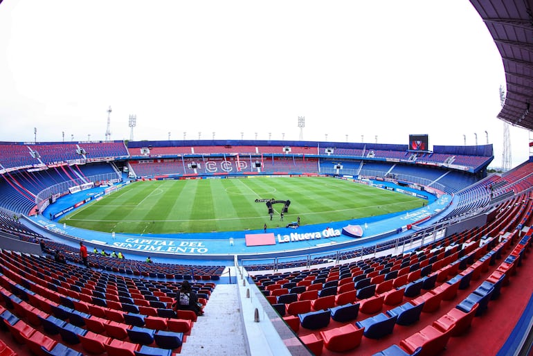 Estadio La Nueva Olla con césped verde y asientos rojos y azules vacíos, banderas y letreros visibles en un cielo nublado.
