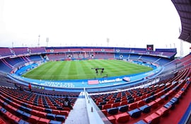 Estadio La Nueva Olla con césped verde y asientos rojos y azules vacíos, banderas y letreros visibles en un cielo nublado.