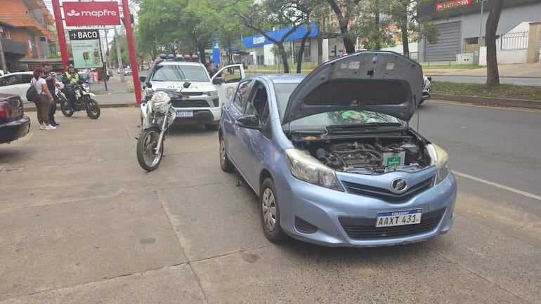 Toyota Vitz, de una empresa de alquiler de vehículos, que tuvo un corte remoto de corriente tras la denuncia de la utilizacion del rodado para robos.
A la derecha de la escena, hay un grupo de tres personas, entre las cuales una está hablando con el oficial, mientras que las otras dos escuchan atentamente. Las personas parecen ser civiles, y no se identifican como figuras públicas reconocibles.
El entorno es urbano, con árboles y edificios visibles al fondo. Destacan algunos carteles publicitarios, pero no hay texto claramente legible. No se observan elementos relacionados con equipos de fútbol ni lugares icónicos. La situación parece ser una reflexión de un incidente o problema vehicular, con la intervención de la policía.