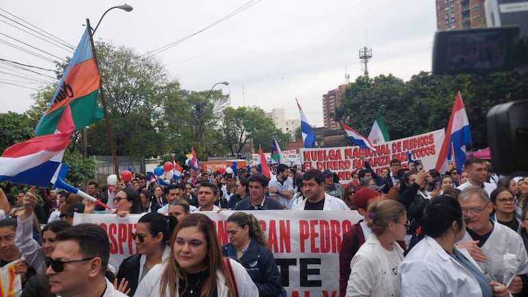 Multitud de médicos en batas blancas sosteniendo una pancarta sobre una manifestación en un entorno urbano con banderas paraguayas.