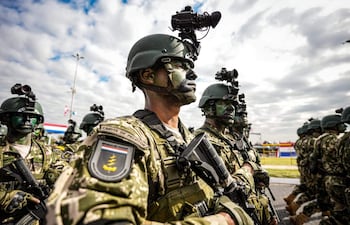 Cinco soldados en fila lucen uniformes de camuflaje, con rostro pintado y casco militar, durante un desfile al aire libre.