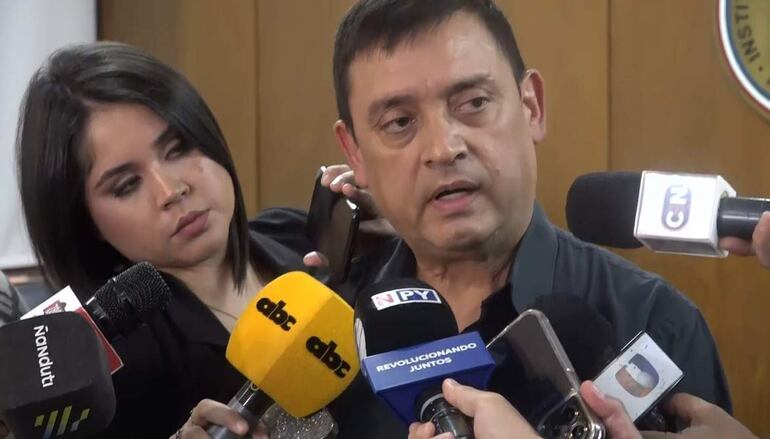 Hombre de camisa oscura habla con expresión seria, mientras mujer a su lado escucha atentamente en sala de prensa.