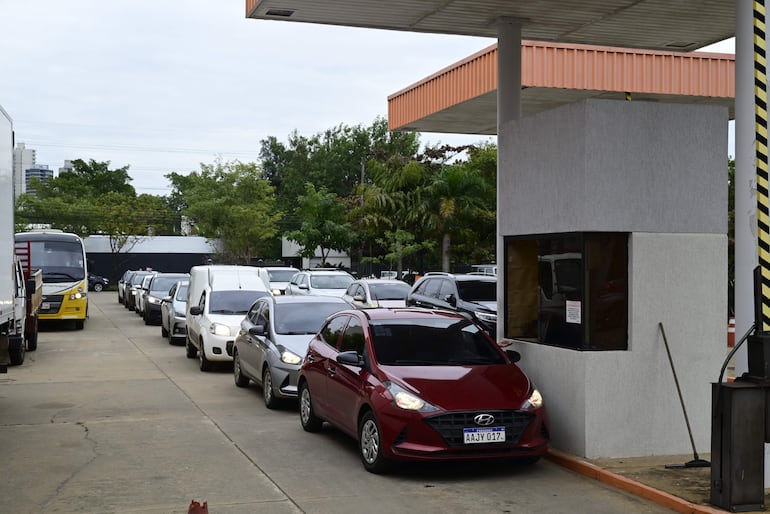 Fila de coches en peaje, predominan automóviles plateados y blancos, con cielo nublado y vegetación al fondo.