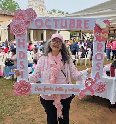 Mujer con cabello rizado y gris, sosteniendo marco con inscripción OCTUBRE ROSA, vestida de rosa en ambiente festivo y solidario.