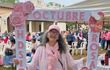 Mujer con cabello rizado y gris, sosteniendo marco con inscripción OCTUBRE ROSA, vestida de rosa en ambiente festivo y solidario.