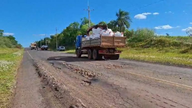 Camión de carga azul transitando por carretera con baches, rodeado de vegetación y cielo despejado.