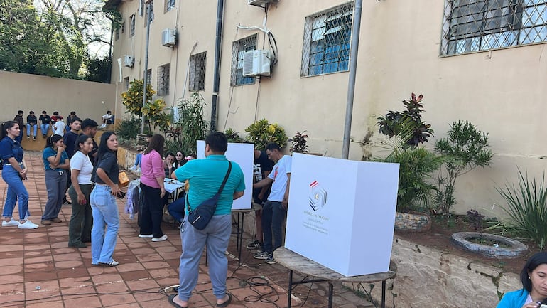 Mujer de pie con un bolso junto a hombres en un ambiente electoral, rodeados de plantas y carteles de la Justicia Electoral.