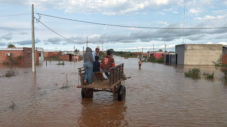 Cuatro personas, tres hombres y un niño, viajan en una carreta de madera en un área inundada, rodeados de vegetación y casas sumergidas.