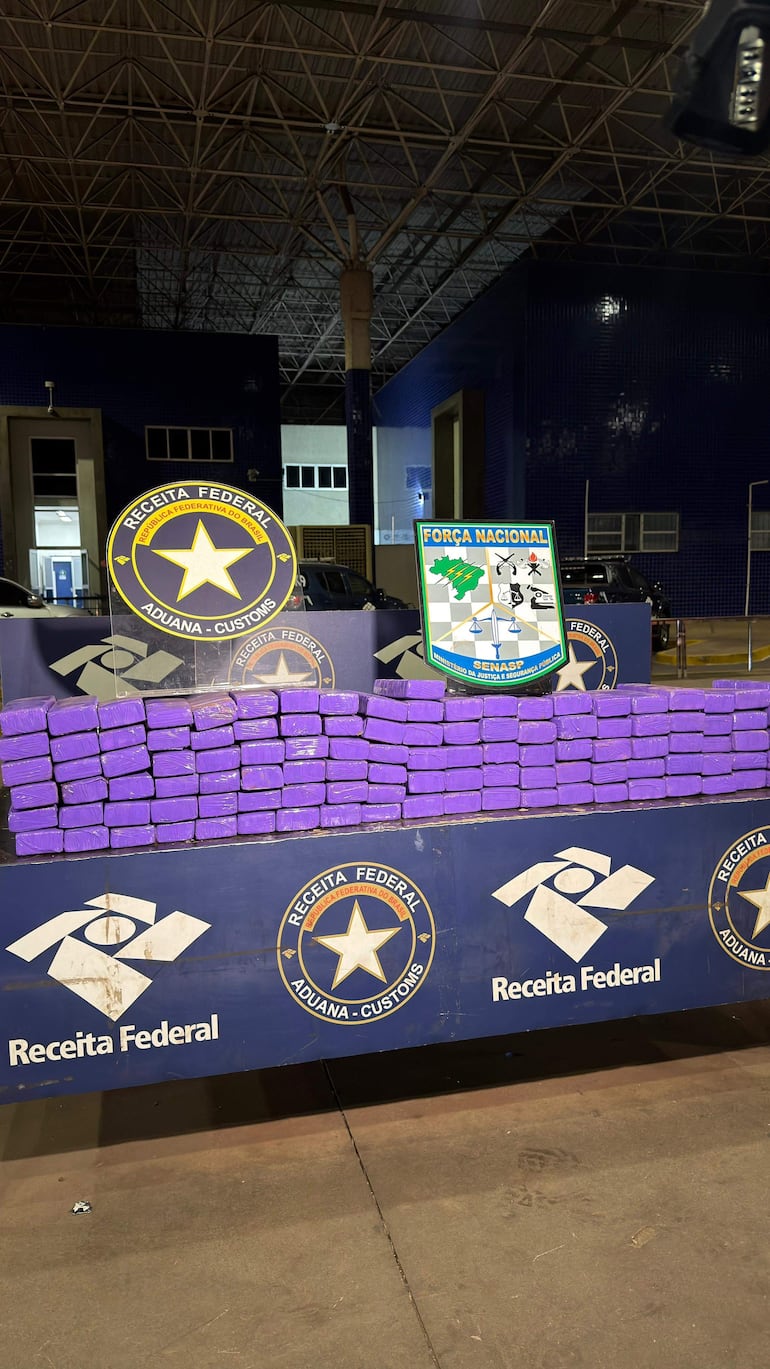 Bloques morados de marihuana confiscados en mesa dentro de una sala aduanera, con logos de Receita Federal y Força Nacional en el fondo.