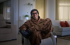 Ronaldinho en sillón blanco, sonriendo y relajado, vestido con sudadera marrón y gorro oscuro en un salón moderno.