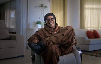 Ronaldinho en sillón blanco, sonriendo y relajado, vestido con sudadera marrón y gorro oscuro en un salón moderno.