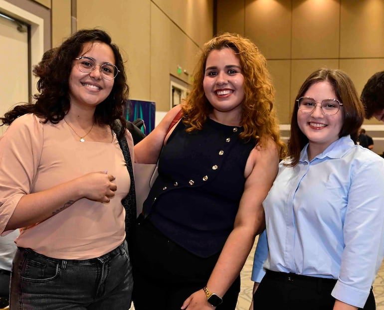 Tres mujeres sonrientes: una con blusa clara y jeans oscuros, otra en blusa oscura con botones y la última en camisa azul clara con gafas.