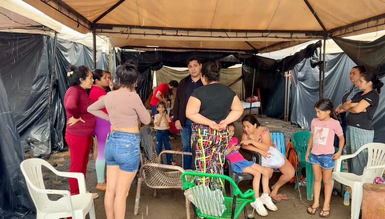 Grupo de seis adultos, la mayoría mujeres, y tres niños conversan bajo un toldo, con un hombre de pie hablando al centro.