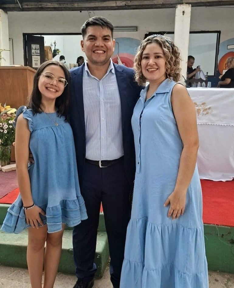 Hombre sonriente en saco oscuro, mujer con vestido de mezclilla y niña con gafas en vestido de mezclilla, todos posando juntos.