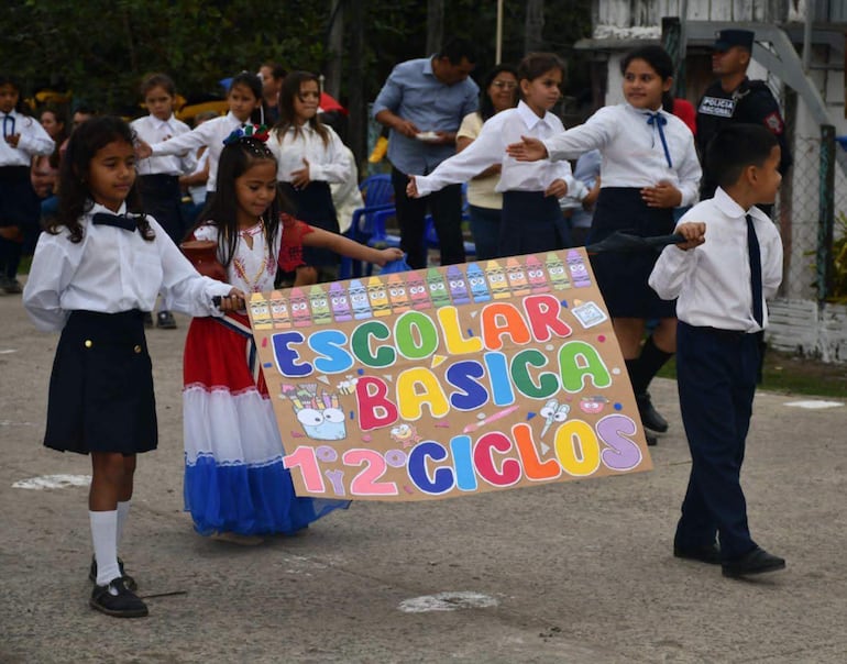 Niños en camisas blancas y pantalones oscuros sostienen un colorido cartel en un ambiente escolar festivo.