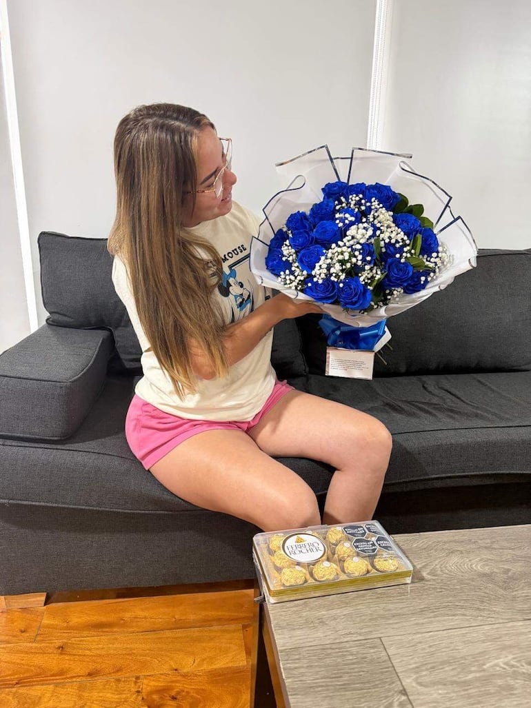 Mujer con cabello largo y lacio, sentada en sofá gris, sosteniendo ramo de flores y observando con alegría.