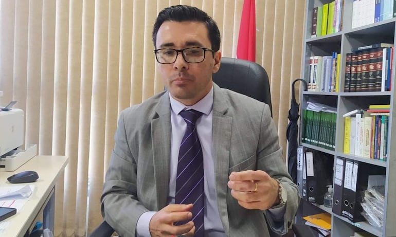 Hombre en traje gris con corbata de rayas, sentado en oficina, gesticulando durante una charla. Fondo con estantería y cortinas beige.